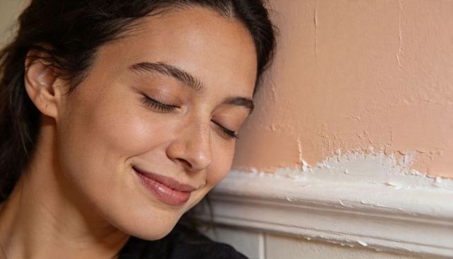 Serene woman resting against peach plaster wall with chipping white trim, intimate closeup