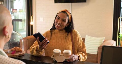 Indian woman ordering takeout coffee at cozy cafe counter while showing phone to barista