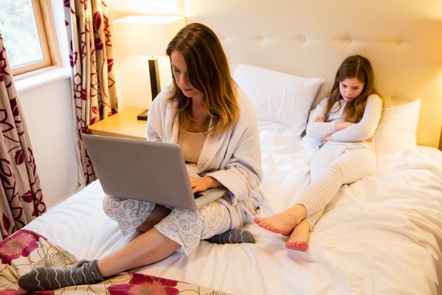 Mother using laptop with her daughter sitting on bed in bedroom