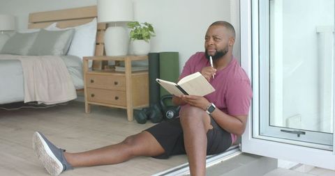 African american man sitting at balcony door journaling and planning home workout routine
