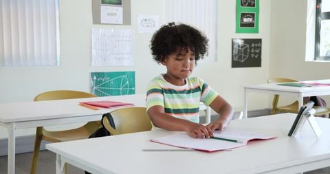 Concentrating African boy studying workbook with tablet and pencil in bright classroom