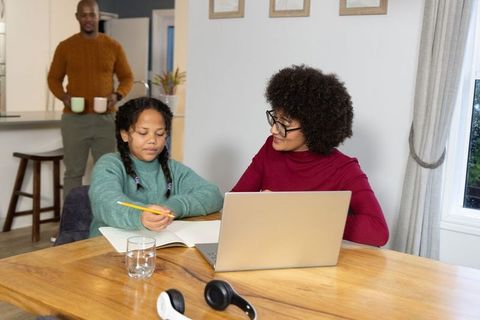 Multiracial parents tutoring daughter at dining table with laptop, father carrying mugs