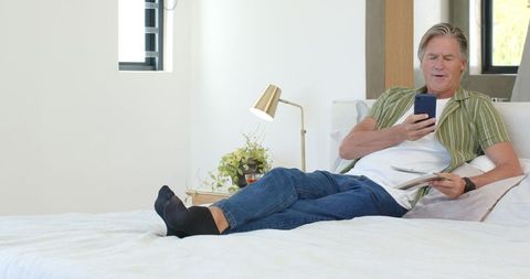 Relaxing senior man using smartphone on bed in bright minimalist bedroom