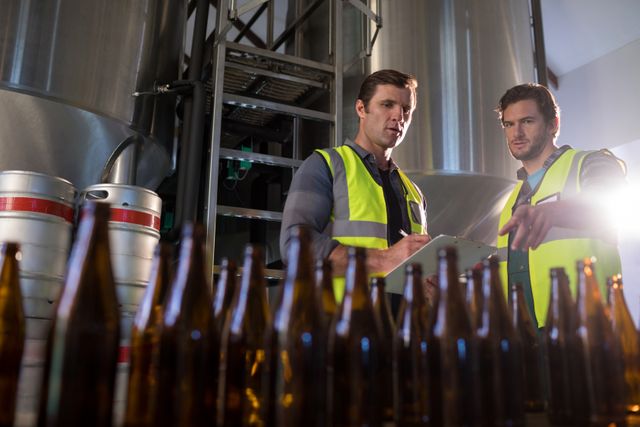 Two coworkers in safety vests discussing beer production process at a brewery. They are pointing towards beer bottles, indicating a focus on quality control and teamwork. This image can be used for articles or advertisements related to the beverage industry, manufacturing processes, workplace safety, and team collaboration.