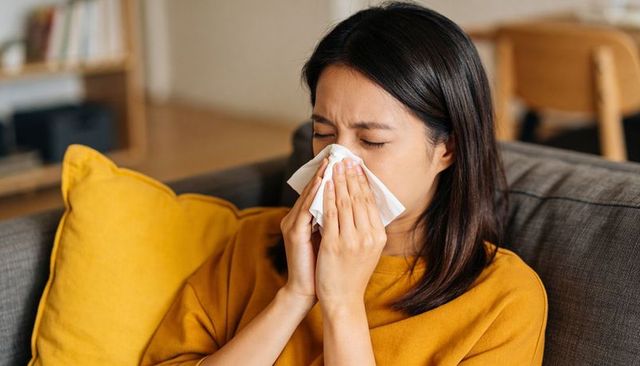 Asian woman sneezing into tissue on cozy sofa at home, seasonal allergy illness concept