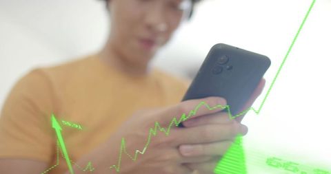 Asian man holding smartphone viewing rising green financial data overlay with stock graph