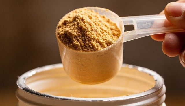 Heaping translucent scoop holding beige powder over open jar close-up for supplement use