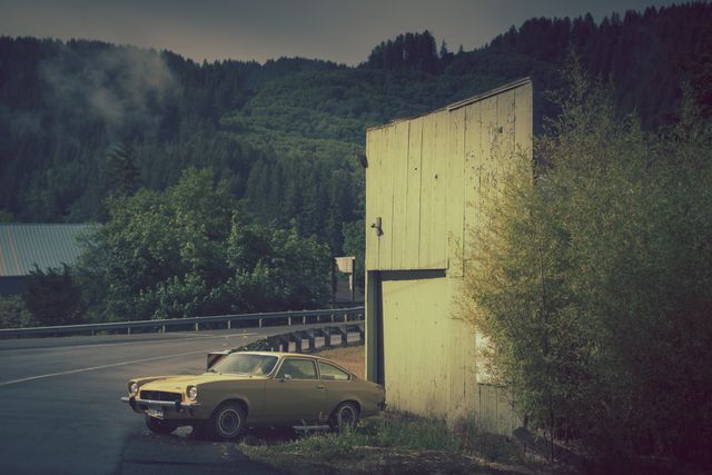 Old yellow car parked next to a weathered building surrounded by greenery and forested hills. Mist rolls in over the mountains, adding a nostalgic feel. Perfect for themes involving history, travel, adventure, and rural atmospheres.