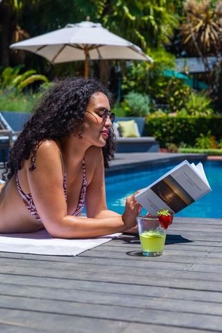 African woman sunbathing and reading paperback by tropical pool with refreshing cocktail