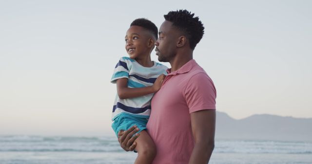 Father holding young son by the beach with ocean and mountains in the background during sunset. Ideal for themes like family bonding, summer vacations, outdoor activities, and heartfelt moments.