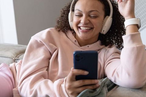 African american woman lounging on sofa smiling, listening with headphones and using smartphone