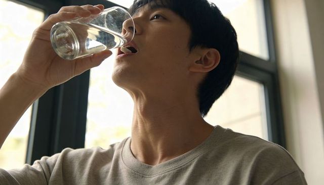 Asian male drinking water by sunlit window, hydration and wellness lifestyle portrait