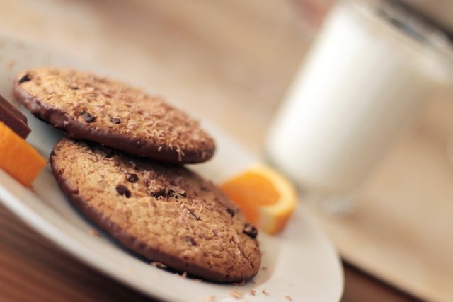 Two chocolate chip cookies on a white plate with orange slices and a glass of milk in the background. Ideal for illustrating concepts of snacks, desserts, and home-baked goods. Perfect for recipes, food blogs, bakery promotions, or lifestyle cooking content.