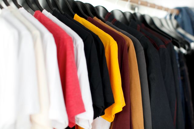 This image depicts a colorful assortment of t-shirts hanging neatly on a rack in a closet. Colors range from white, red, yellow, to various shades. This can be used to depict fashion trends, casual apparel, wardrobe organization, and retail clothing displays.