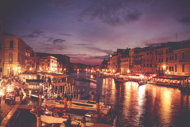 Illuminated Venetian canal during twilight featuring vibrant nightlife along the water, with gondolas floating. Historic buildings reflecting in the water give a sense of the romantic, cultural destination. Ideal for travel advertisements, brochures, and romantic getaway promotions showcasing Venice at night.