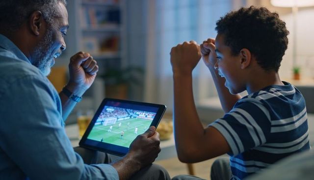Senior man and teen boy cheering while watching live soccer on tablet in cozy living room