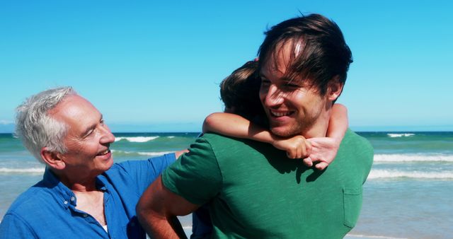 A middle-aged man and a senior man enjoy a warm embrace on a sunny beach, with the ocean in the background. Their joyful interaction captures a moment of family connection and affection against a beautiful coastal backdrop.