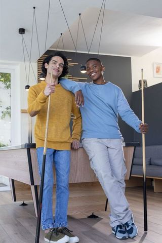 Multiracial adolescent male friends leaning on pool table in modern game lounge, smiling