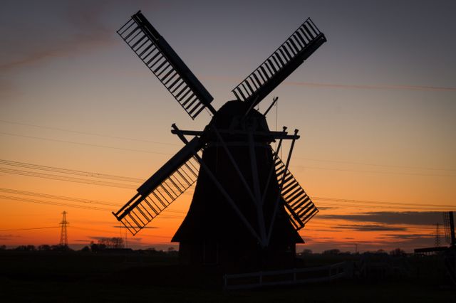 Silhouetted windmill standing against a vibrant sunrise in a rural landscape. Suitable for use in travel blogs, promotional materials for countryside tourism, or portraying picturesque rural life. Great for backgrounds or inspiring quotes about tranquility and nature.