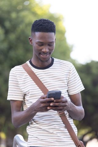 Young black man texting and smiling while sitting on park bench with crossbody bag
