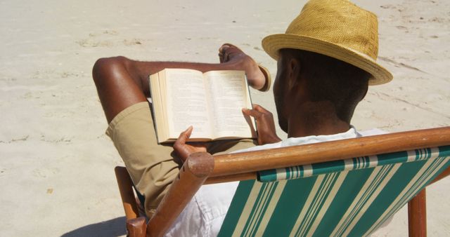 An African American individual relaxes on a beach chair, engrossed in reading a book, with copy space. A straw hat and sunny beach setting suggest a leisurely vacation or a peaceful retreat.