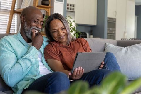 Mature african american couple sitting on sofa, smiling and using tablet at home, cozy