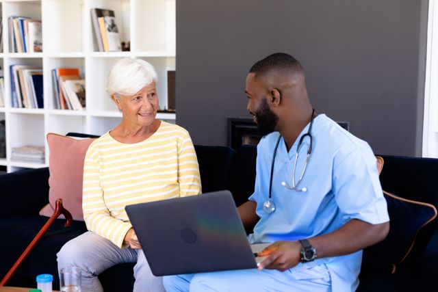 African american male doctor holding laptop talking with biracial senior woman while sitting on sofa. Home, technology, unaltered, physical therapy, healthcare, patient, retirement and recovery.