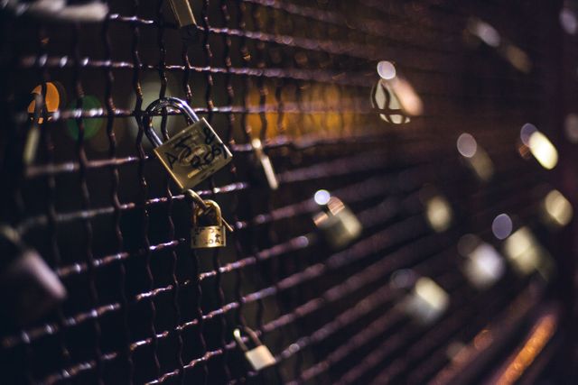 Close-up of multiple padlocks attached to a chain-link fence, commonly referred to as love locks. Captured in an urban setting at night, the image features a shallow depth of field with bokeh lights in the background. Suitable for themes of commitment, love, security, and urban life.