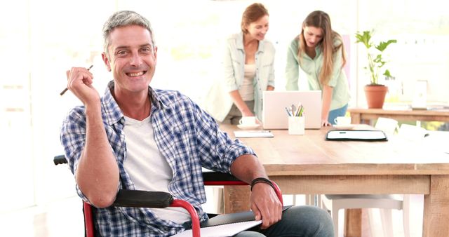 Businessman in a wheelchair happily working with colleagues in a modern office environment. A laptop, notebooks, and a cup are on the table, indicating an active work meeting. This image can be used to represent inclusivity, teamwork, and a positive workplace attitude. Ideal for showcasing diversity in professional settings, promoting inclusive work environments, and illustrating collaborative business efforts.