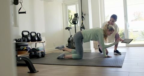 Personal trainer guiding client through kneeling plank in bright home gym using tablet
