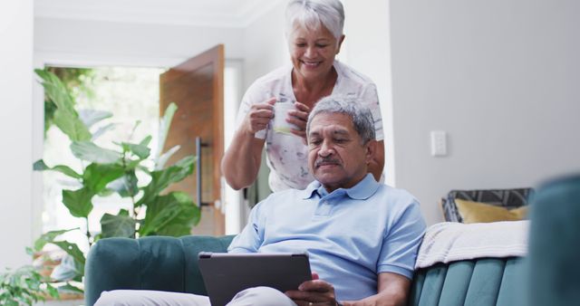 Happy senior biracial couple with coffee using tablet. Spending quality time at home, retirement and lifestyle concept.