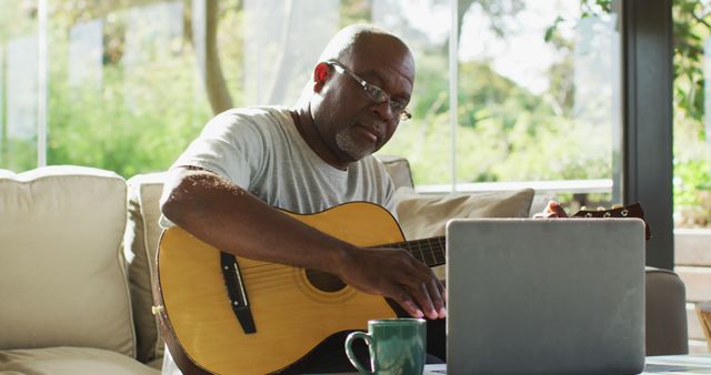 Older African American man playing guitar while engaging with laptop at home. Perfect for concepts related to remote learning, online guitar lessons, hobbies, leisure activities, and enjoying retirement. Ideal for educational websites, music and hobby-related content, or lifestyle blogs.