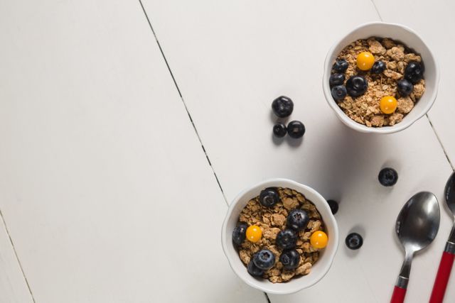 Two bowls filled with granola, blueberries, and yellow berries on white background. Spoons and scattered berries add to the composition. Ideal for promoting healthy eating, breakfast ideas, or nutrition-focused content.