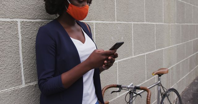 Young woman standing against wall using smartphone while her bicycle is parked beside her. Face mask suggests precautions during covid-19 pandemic. Suitable for topics related to safety, current events, urban lifestyle, technology, communication, and casual outdoor settings.