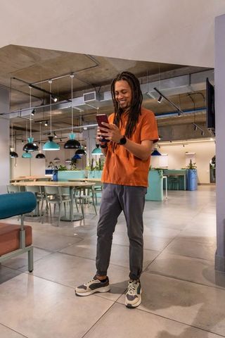 Young African American man standing in modern coworking space using smartphone and smiling