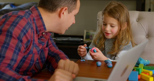 Father engaging with daughter in a creative activity with modeling clay in a cozy home environment. Can be used for stories on parenting, family bonding, indoor activities for children, and arts and crafts. Perfect for illustrating parent-child interactions, creative playtime, and family moments.