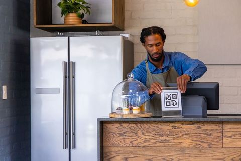 African american barista placing qr code sign at wooden counter with cupcakes