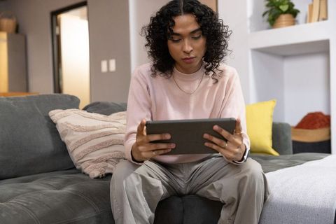 Non-binary person relaxing on sofa using tablet and smartwatch in modern cozy living room