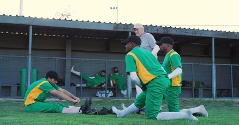 Male baseball team warming and stretching on grass beside dugout with coach observing