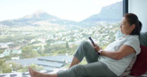Senior asian woman reclining by panoramic window using smartphone and gazing over valley