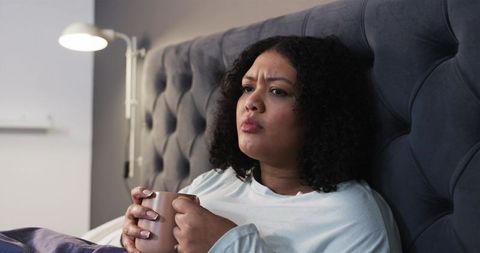 African American woman sitting in bed holding mug looking pensive beside tufted headboard