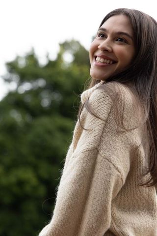 Smiling woman looking up in park wearing cozy beige knit sweater with natural bokeh greenery