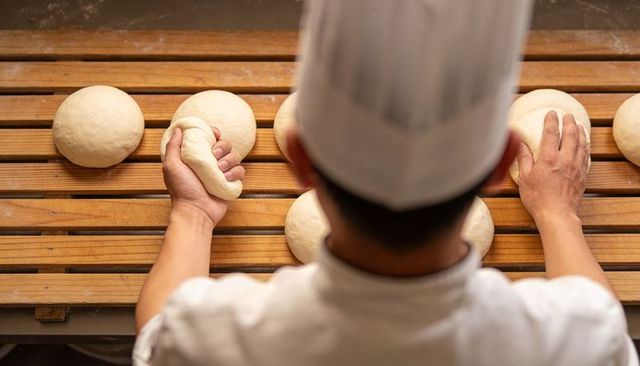 Baker shaping dough balls on wooden proofing board, artisan kneading hands at work in bakery