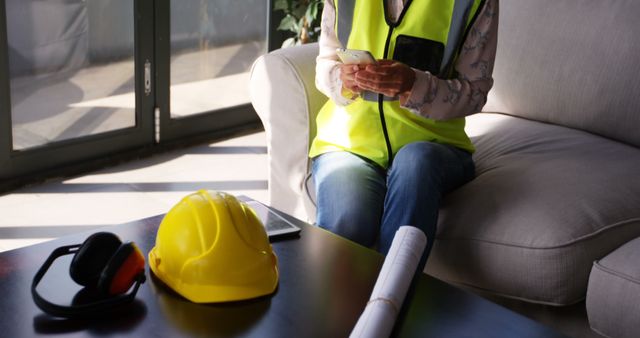 Architect, dressed casually with a safety vest, sitting on a couch using a smartphone. A hard hat, ear protection, and blueprints are on the table in front of her. Ideal for professional workplace, construction, architectural industry, or safety gear usage imagery.