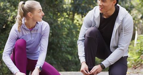 Kneeling fitness partners tying running shoes on park trail wearing smartwatches