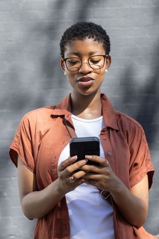 African woman using smartphone in sunlit urban scene wearing glasses and rust shirt