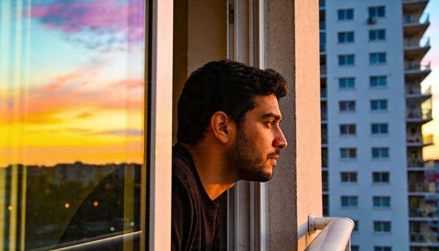 Young man leaning on balcony watching colorful sunset over urban skyline with window reflection