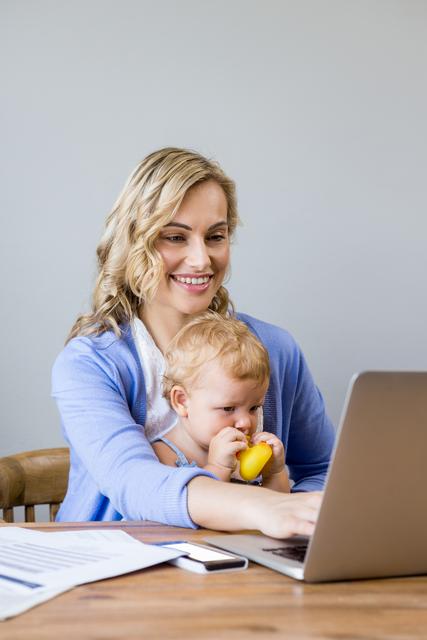 Mother working on laptop while holding baby at home. Ideal for illustrating concepts of remote work, multitasking, parenting, work-life balance, and family life. Suitable for articles, blogs, and advertisements related to working mothers, home office setups, and family dynamics.