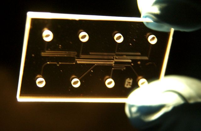 Microfluidic chip held by gloved hands, designed by NASA's Marshall Space Flight Center for growing biological crystals on the International Space Station. The chip features eight ports that can be filled with fluids or chemicals for diverse applications such as medical tests, water quality monitoring, and detecting life signatures on other planets. This image can be used in content related to space research, biotechnological advancements, and educational materials about scientific experiments in space.