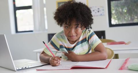 Curly-haired student writing in notebook with laptop at classroom desk, focused learning